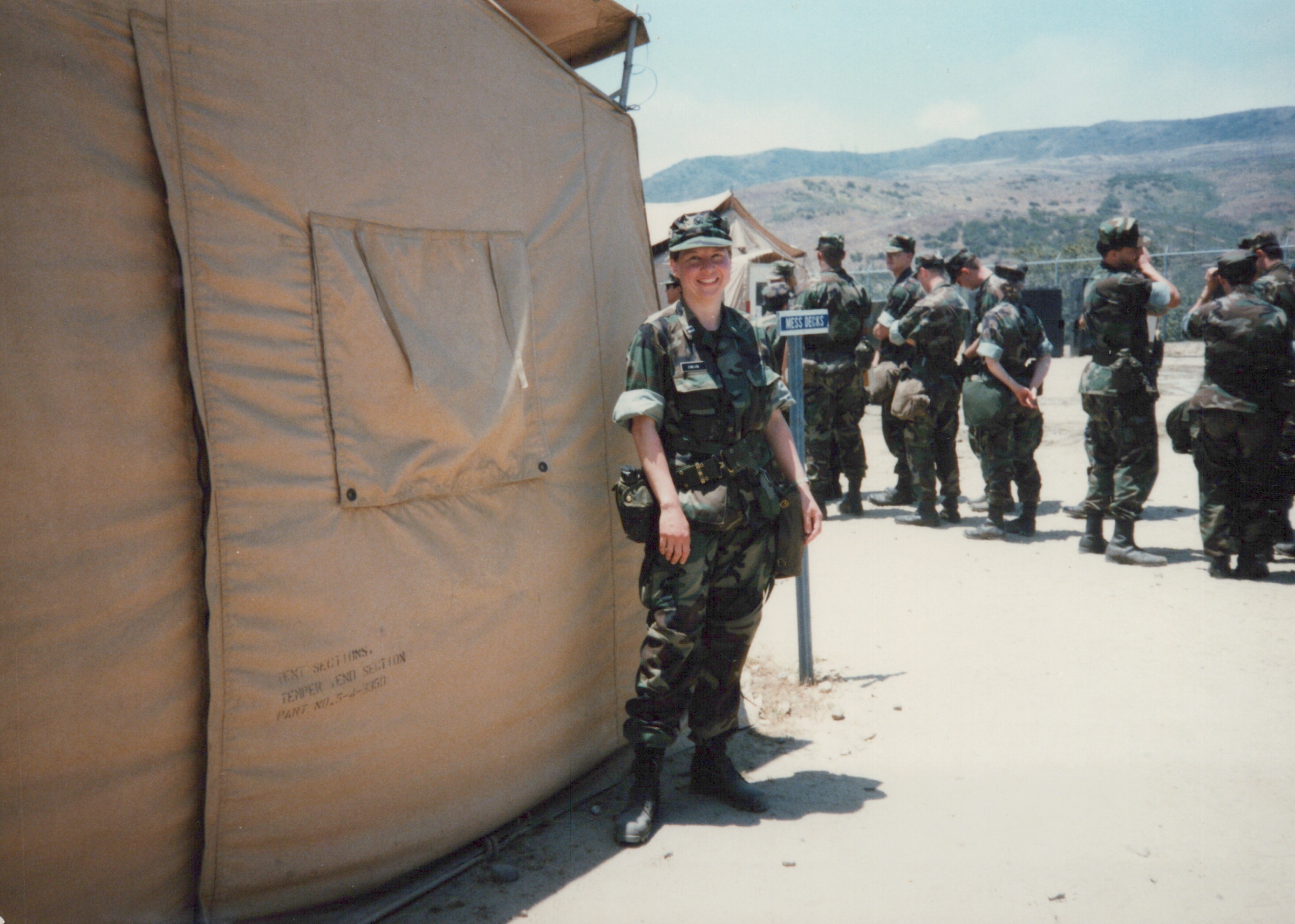 Fleet Hospital 007: Uniformed person wearing a side pouch stands beside a tent; others gather behind.